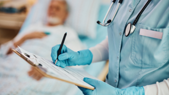 Torso de mujer con pijama sanitario azul tomando notas al lado de una cama con paciente desenfocada