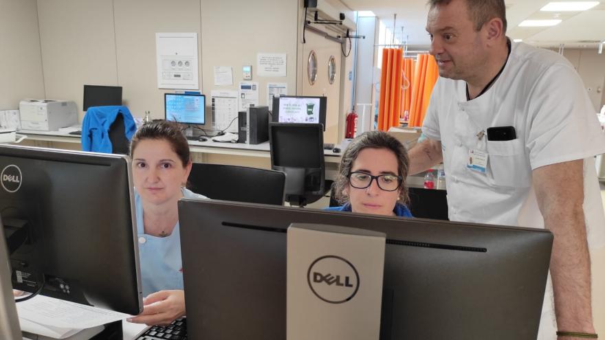 Dos mujeres sentadas delante del ordenador junto a un hombre de pie en el interior de una sala de Urgencias