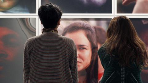CA2M Centro de Arte Dos de Mayo Dos mujeres sentadas de espaldas al espectador y mirando una pared llena de fotografías con retratos en color de personas anónimas