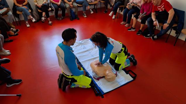 Dos profesionales del SUMMA 112 se encuentran de rodillas frente a un muñeco de formación realizando las técnicas de reanimación cardiopulmonar rodeado de un grupo de ciudadanos que se encuentran formándose en técnicas de reanimación 