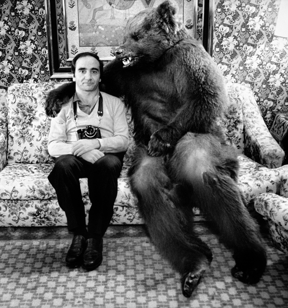 Man sitting on a sofa next to a brown bear that hugs him with its paw on his shoulder