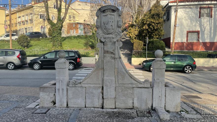 Front image of a stone fountain with two side columns, a central pillar with a shield and inscriptions