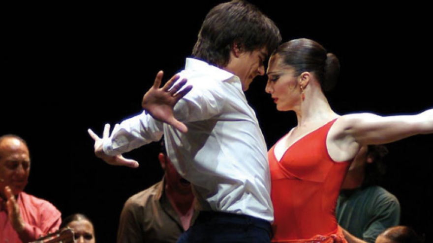 Dos bailarines abriendo los brazos, parte superior del cuerpo, él con camisa blanca ella con vestido rojo