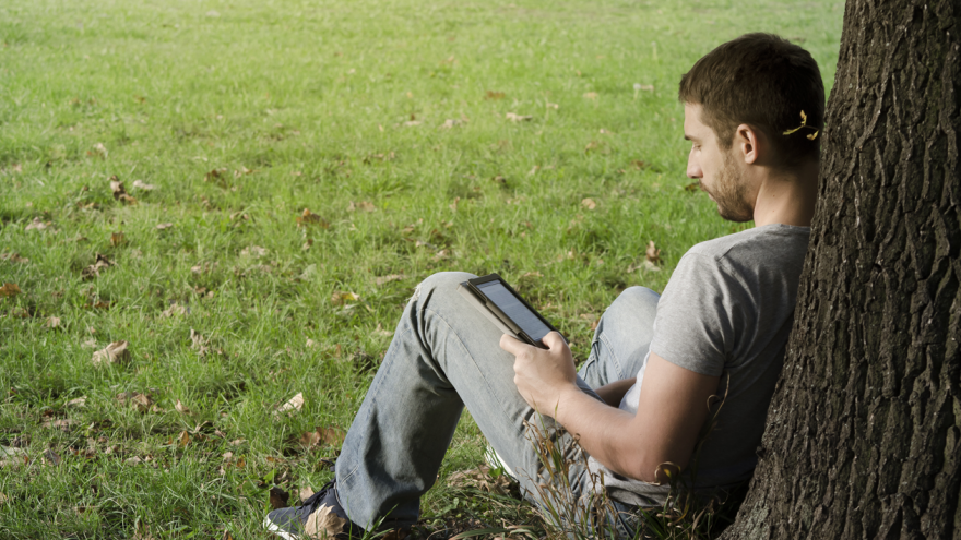 Chico leyendo en tablet apoyado en un árbol