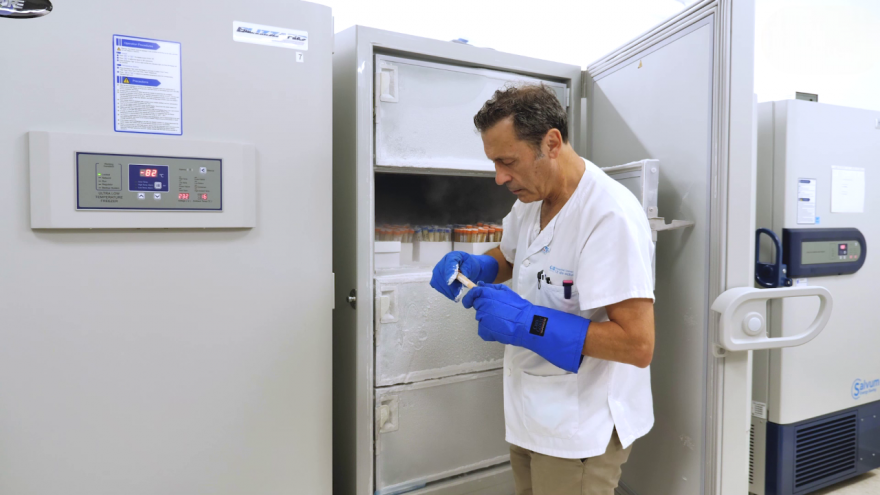 profesional con chaquetilla blanca y guantes azules sacando tubos de un congelador de laboratorio