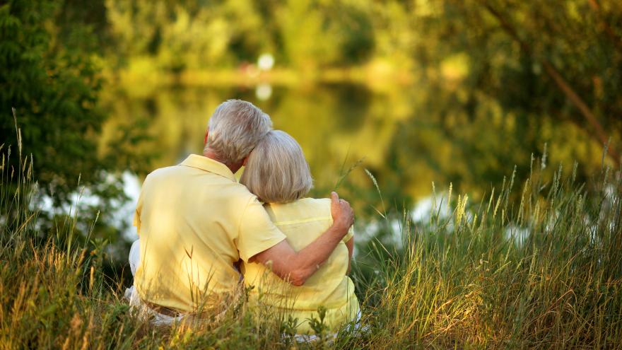 Dos personas mayores abrazadas sentadas a los pies de un lago