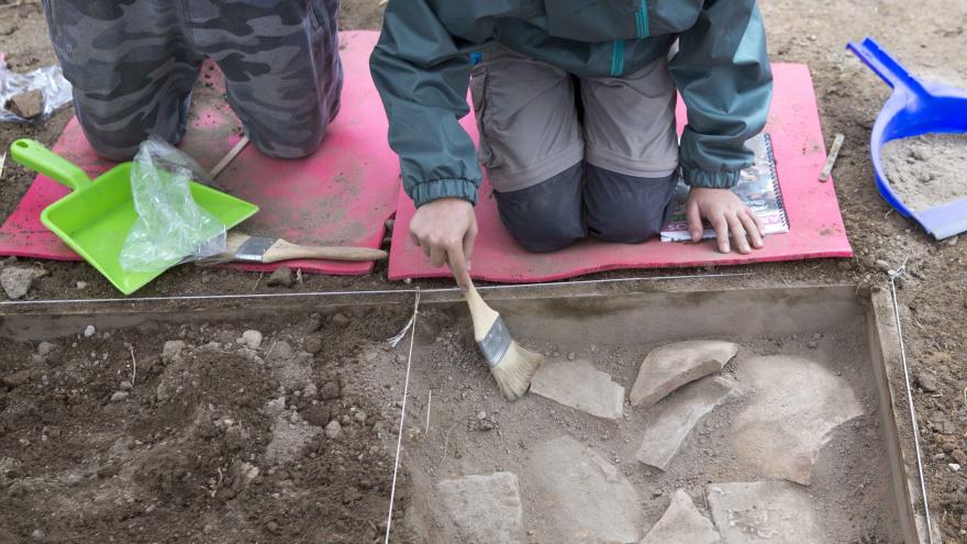 Niños arrodillados en una imitación de yacimiento arqueológico limpiando restos de cerámicas con unas brochas