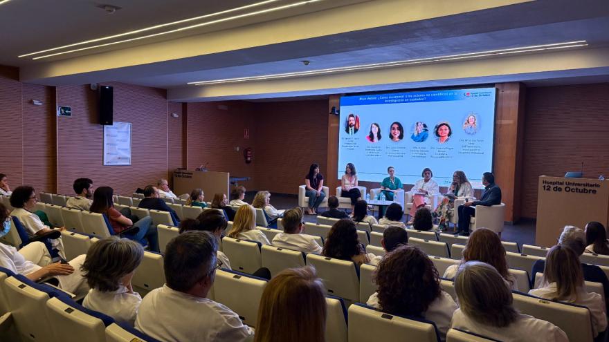 Salon de actos visto desde atrás lleno y en escenario varios sillones con personas sentadas y pantallas detrás