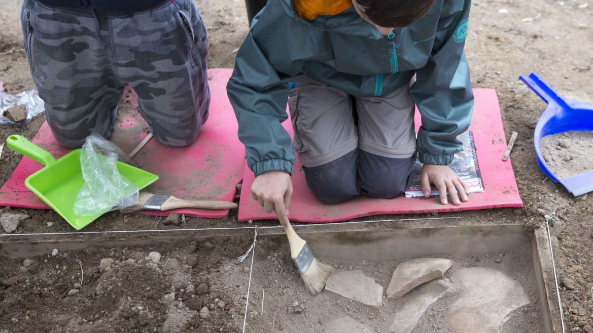 Niños arrodillados en una imitación de yacimiento arqueológico limpiando restos de cerámicas con unas brochas