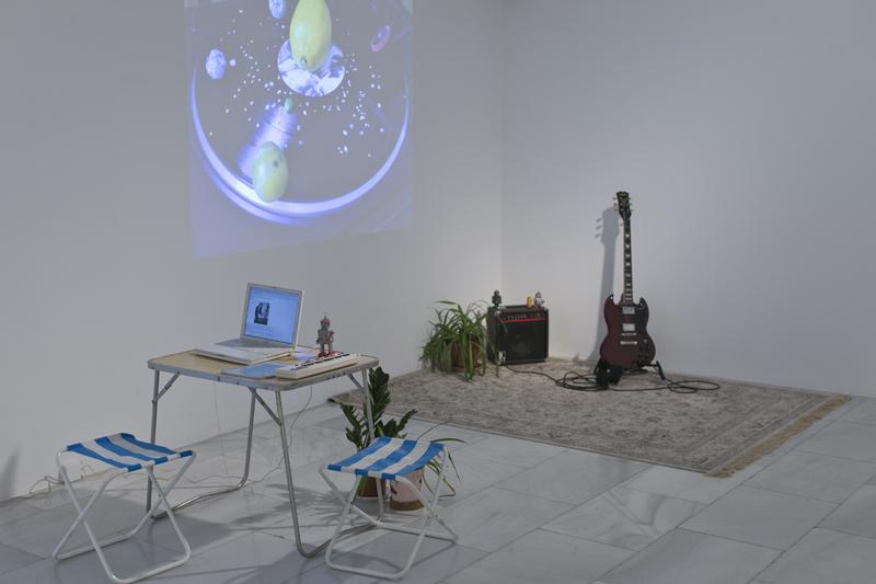 Artistic installation with various pieces such as a table, a computer, a carpet, an electric guitar