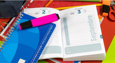 Sobre una mesa de escritorio hay una agenda, un cuaderno y un marcador fluorescente