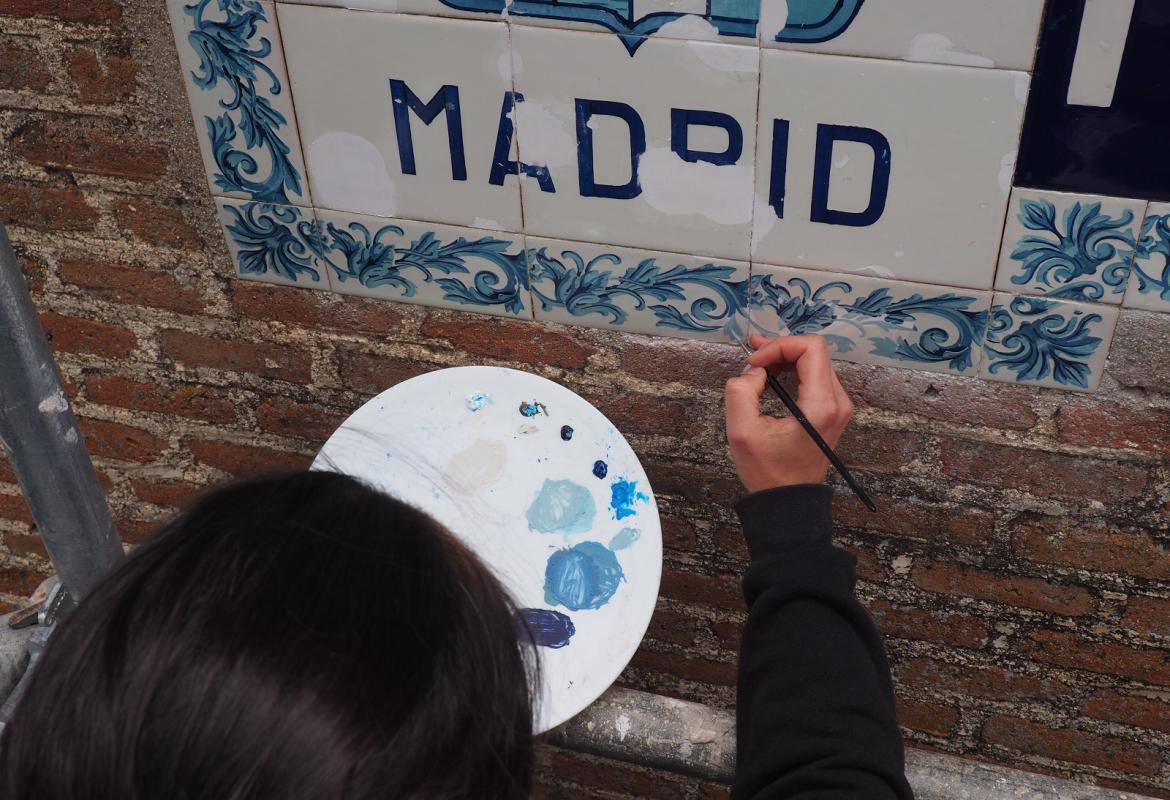 paleta de pintura con tonos azules en primer plano, una mano con pincel reintegra las partes perdidas de un escudo de azulejos